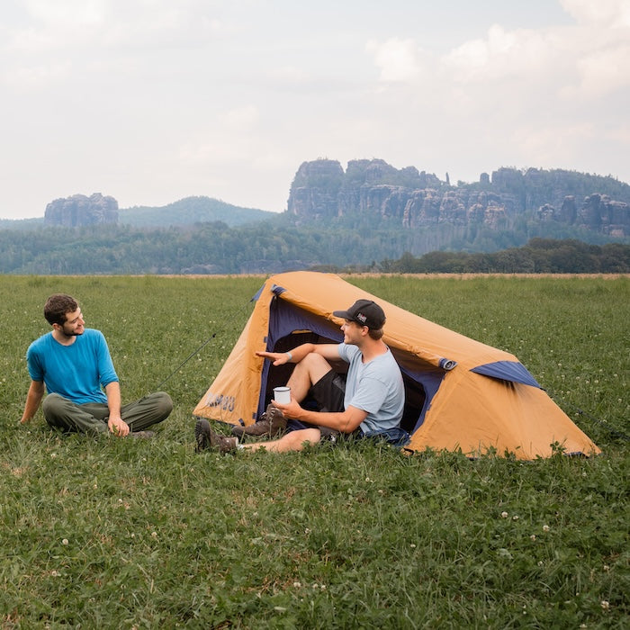 Zwei Männer sitzen auf einer Wiese vor einem gelbfarbenen SURANGA 2.0 Leichtgewichts-Trekkingzelt von Tambu Outdoor, im Hintergrund Felsenlandschaft