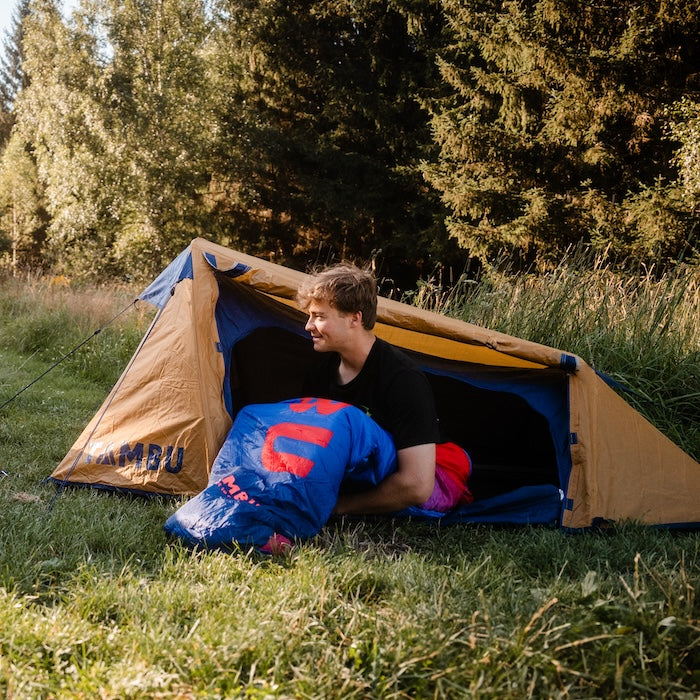 Mann sitzt am Eingang des gelb-blauen SURANGA 2.0 Leichtgewichts-Trekkingzelts von Tambu Outdoor und hält blauen Schlafsack