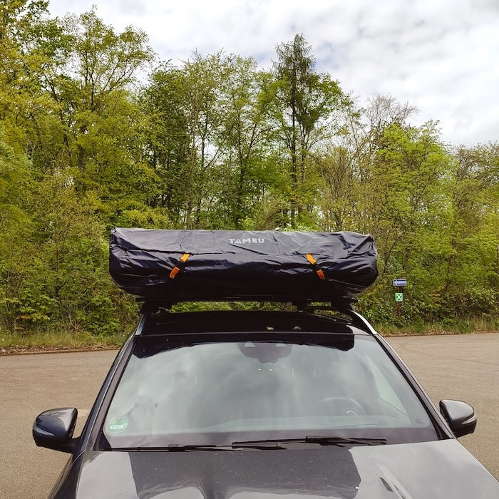 Schwarze YANO Abdeckhaube von Tambu Outdoor auf einem Dachzelt, montiert auf dem Dach eines schwarzen Autos vor bewaldetem Hintergrund