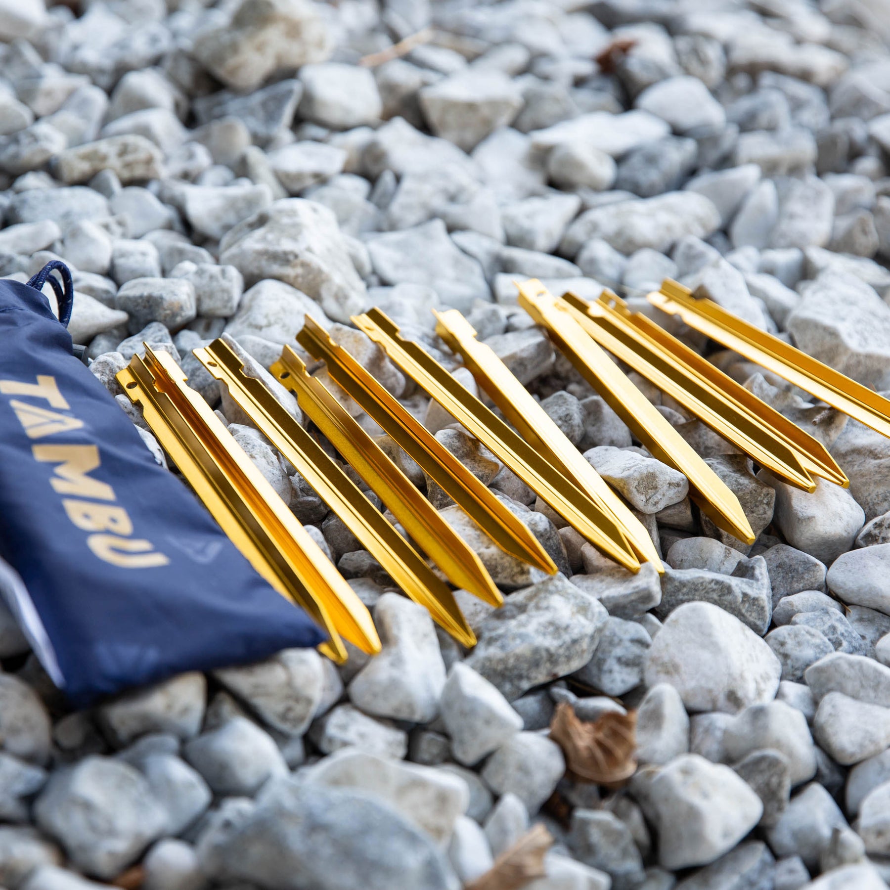 Zwölf ultraleichte goldfarbene Aluminium-Heringe von Tambu Outdoor auf grauem Kies, neben blauem Aufbewahrungsbeutel mit 'TAMBU' Schriftzug
