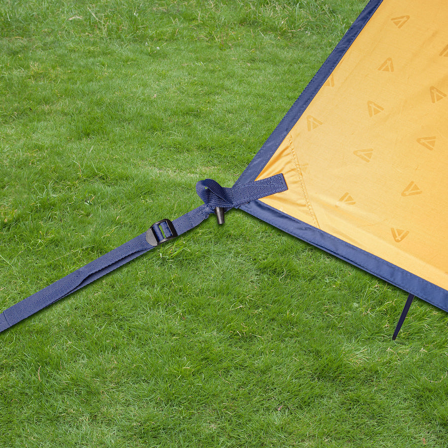 Ecke eines gelben CHADA Sonnensegels von Tambu Outdoor mit dunkelblauem Rand und Befestigungsgurt auf grünem Gras