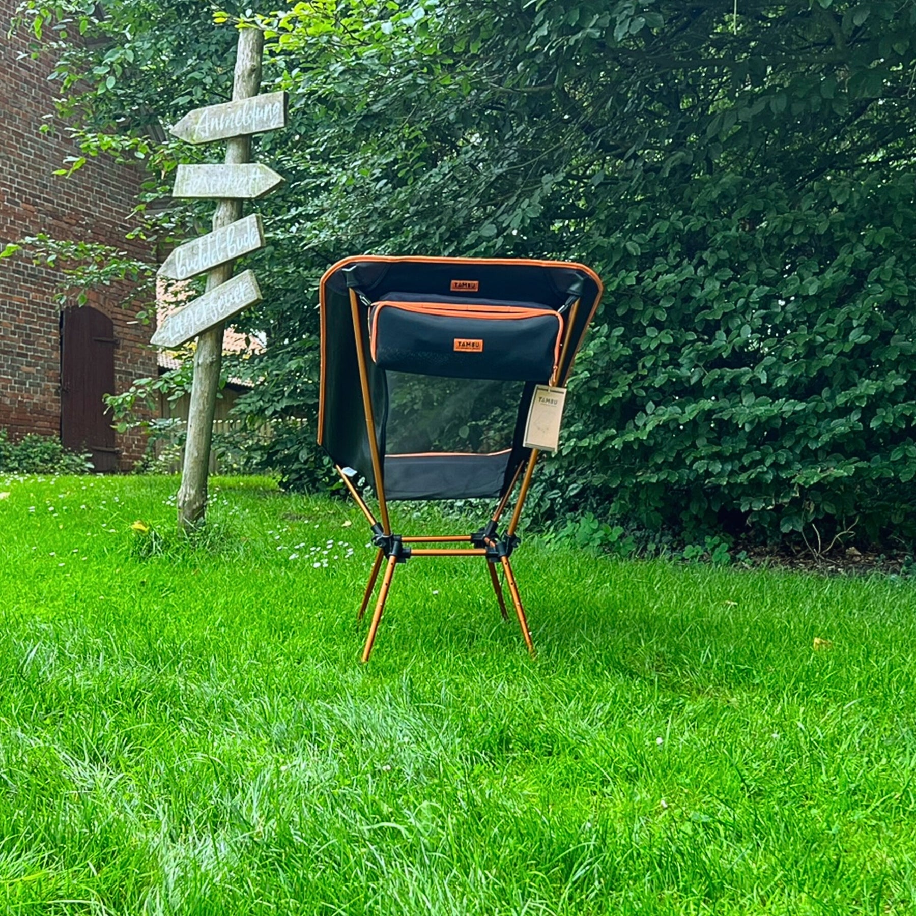 Schwarzer und oranger leichter Campingstuhl BATASA COMFORT von Tambu Outdoor mit vier Sitzhöhen auf grünem Rasen vor Büschen und Holzwegweiser