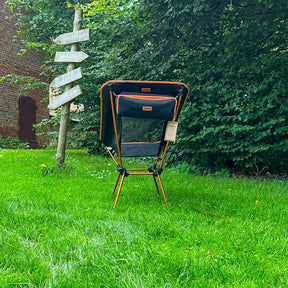 Schwarzer und oranger leichter Campingstuhl BATASA COMFORT von Tambu Outdoor mit vier Sitzhöhen auf grünem Rasen vor Büschen und Holzwegweiser