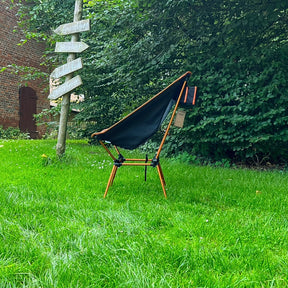 Leichter Campingstuhl BATASA COMFORT von Tambu Outdoor in Schwarz und Orange mit vier Sitzhöhen, auf grünem Rasen vor Büschen und Holzwegweiser