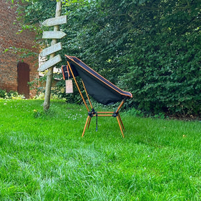 Schwarzer und orangefarbener leichter Campingstuhl BATASA COMFORT von Tambu Outdoor mit vier Sitzhöhen, auf grünem Rasen vor Büschen und einem Holzwegweiser