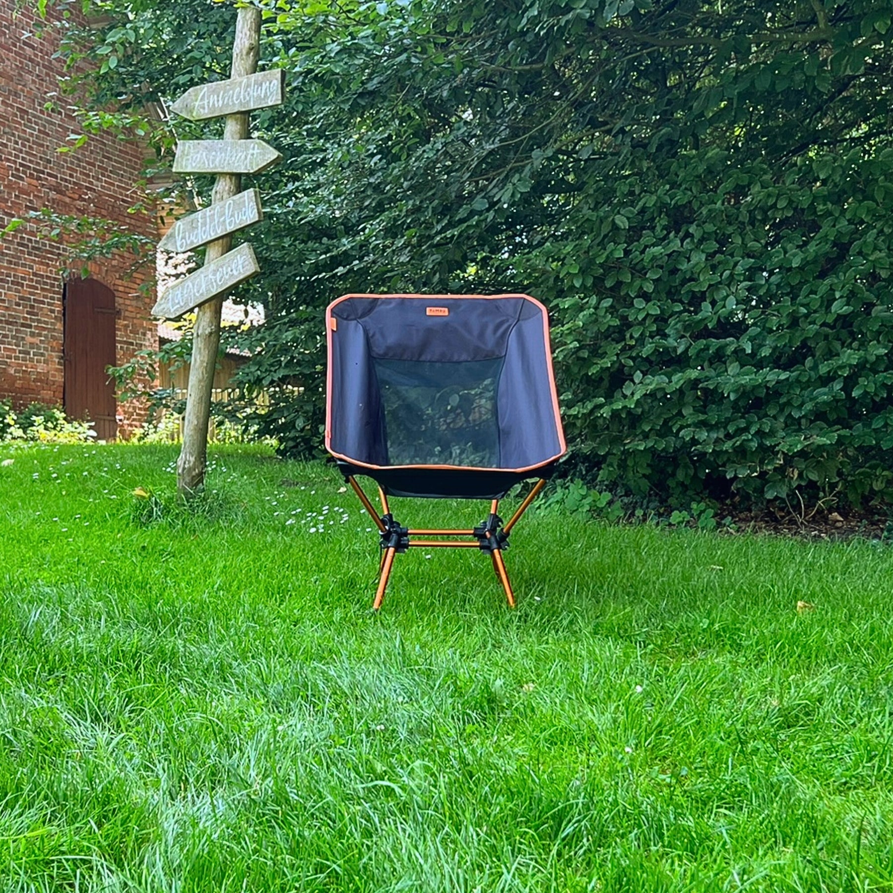 Schwarzer und oranger leichter Campingstuhl BATASA COMFORT von Tambu Outdoor auf grünem Gras vor Büschen und Holzwegweiser