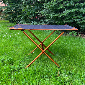 Leichter Campingtisch BATISTA von Tambu Outdoor mit schwarzer Tischplatte und orangefarbenem Gestell auf grünem Gras