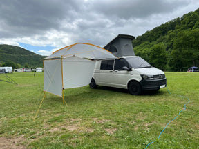 Weißer Van mit aufgestelltem Dachzelt und angebrachtem BASANT multifunktionalem Sonnensegel von Tambu Outdoor auf einer grünen Wiese mit bewaldeten Hügeln im Hintergrund, mit Seitenwände
