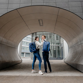 Zwei Personen stehen sich in einem modernen Tunnel gegenüber, beide tragen den wasserabweisenden ANIK 22 oder 28 Rucksack von Tambu Outdoor in Blau.